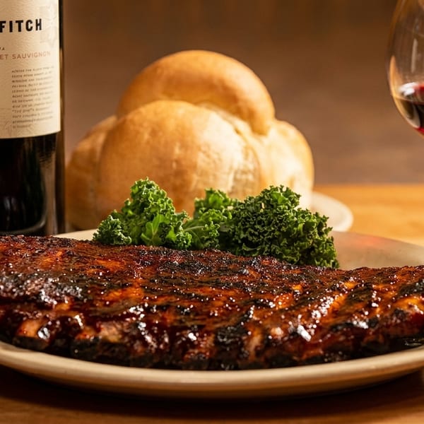 A rack of BBQ ribs with kale, bread rolls, a bottle of red wine, and a glass of wine.