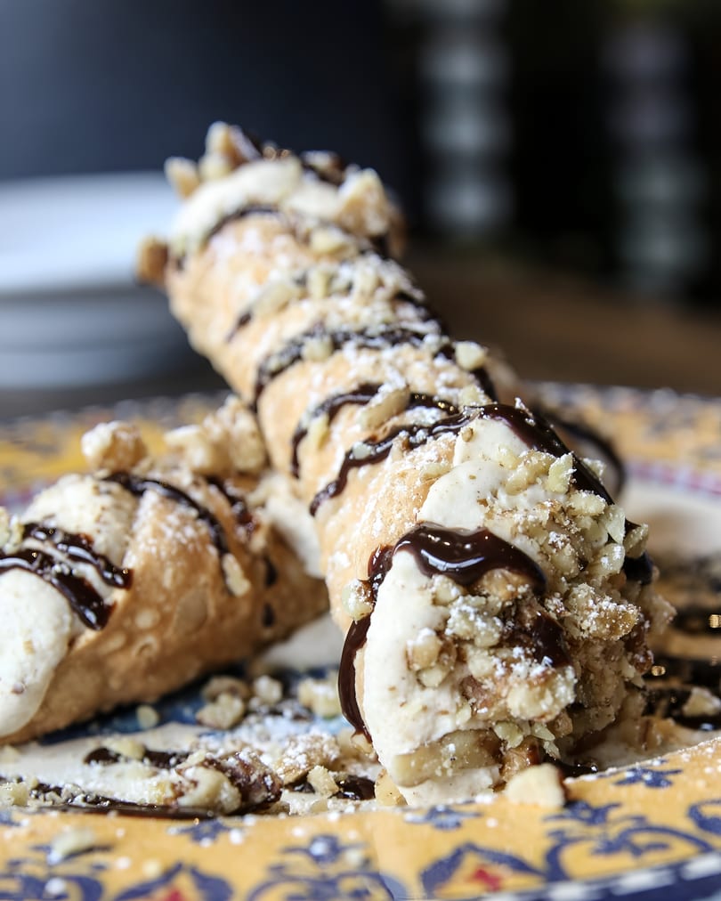 Two cannolis filled with cream, drizzled with chocolate, and topped with nuts and powdered sugar.