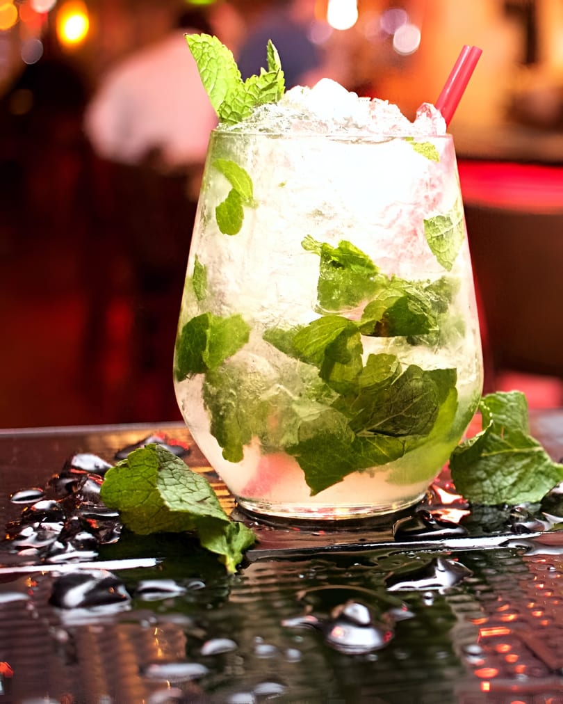 A refreshing mojito cocktail with crushed ice, fresh mint leaves, and a red straw on a wet surface.