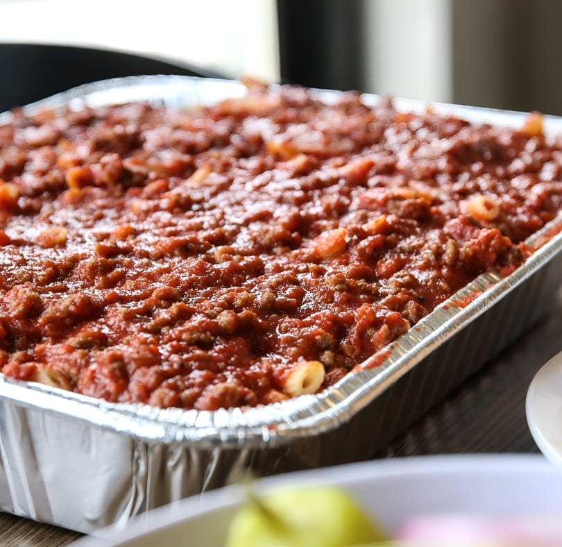 A large aluminum tray filled with baked ziti pasta covered in a rich red meat sauce.