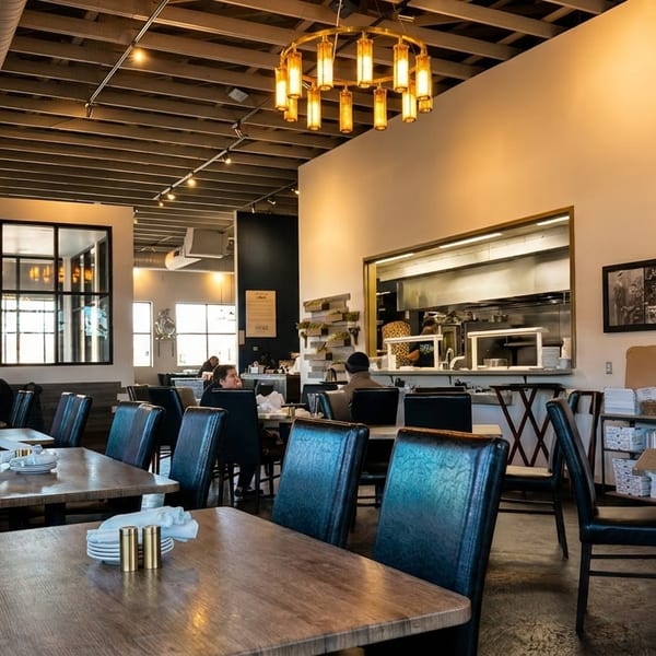 A restaurant interior with patrons dining, featuring a long booth, wooden tables, and an open kitchen.