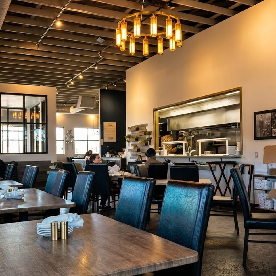 A restaurant interior with patrons dining, featuring a long booth, wooden tables, and an open kitchen.