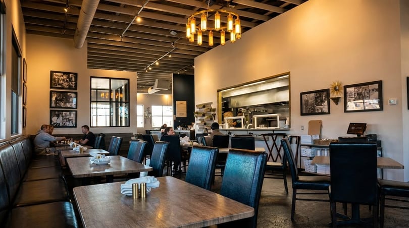 A restaurant interior with patrons dining, featuring a long booth, wooden tables, and an open kitchen.