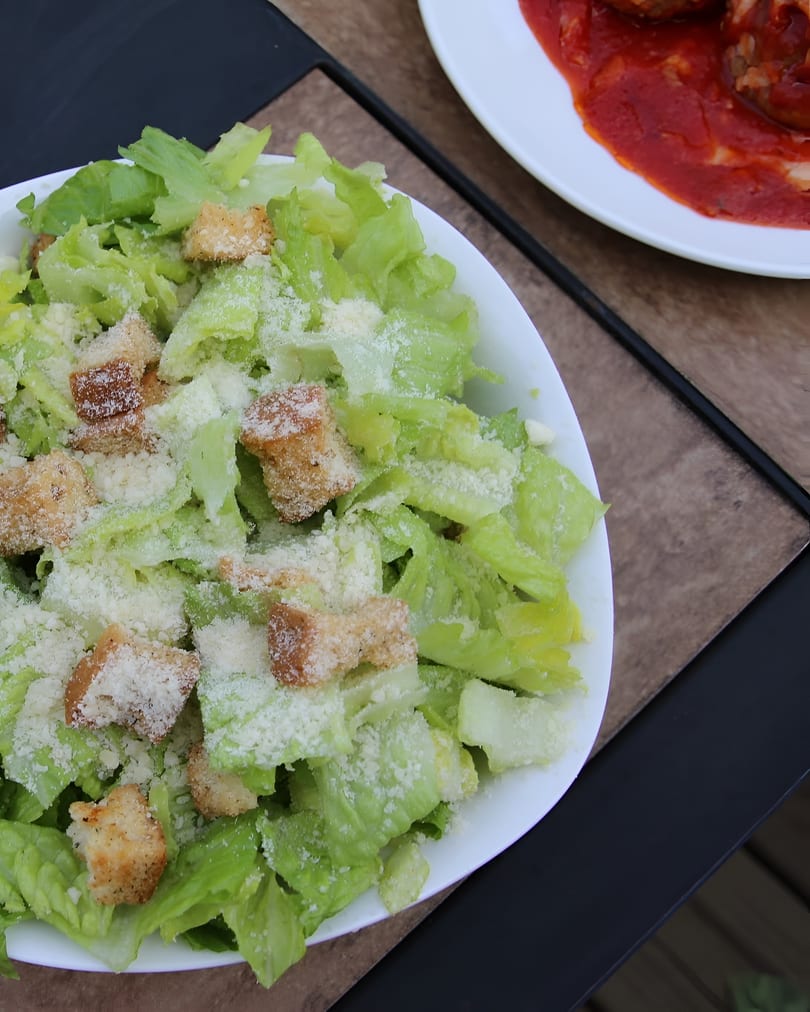 A white bowl of Caesar salad with croutons and parmesan cheese, next to a plate of meatballs.